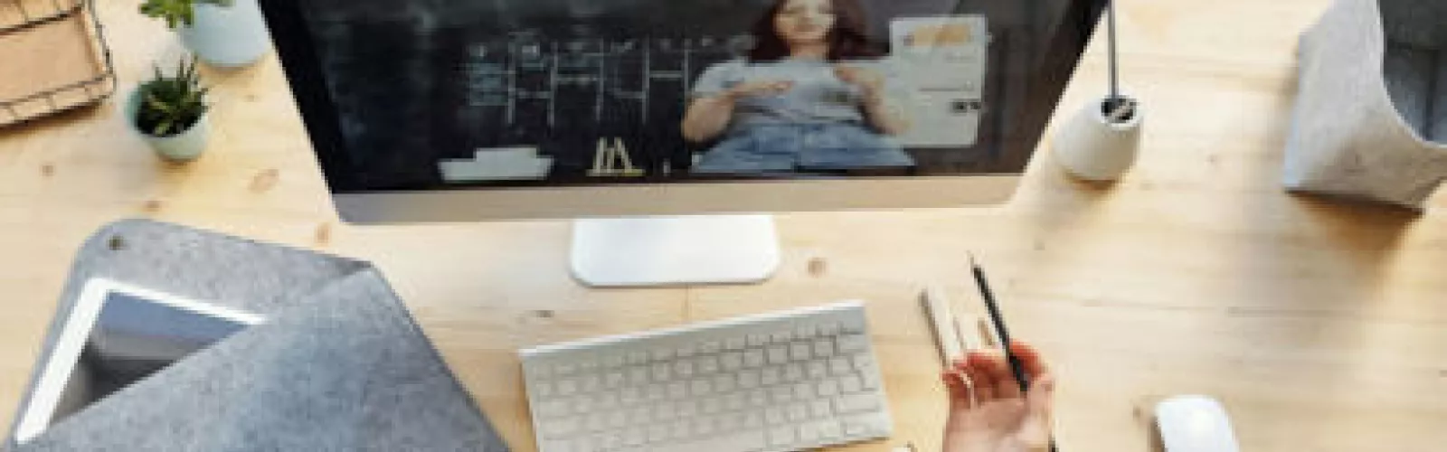 A hand holding a pencil over a desk, facing a computer screen showing  a person with a blackboard
